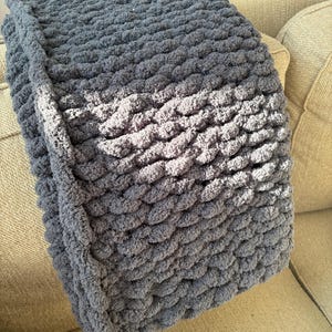 May include: A folded, chunky knit blanket in shades of blue-gray. The blanket has a textured, looped design and is resting on a beige sofa. The soft, plush material is visible in the close-up shot.