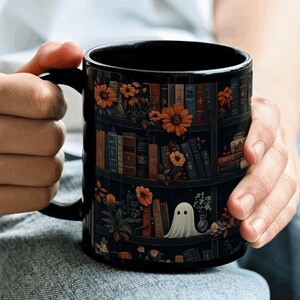 May include: Black ceramic mug featuring a bookshelf design with orange flowers, books, and a ghost illustration. The mug has a black handle and interior. The design wraps around the mug, creating a whimsical and cozy aesthetic.