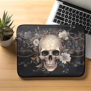 Op de afbeelding: Een zwarte laptophoes met een witte schedel omringd door witte bloemen en gouden kettingen op een donkere achtergrond.