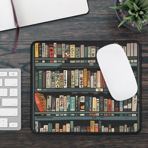 May include: A black mouse mat with a bookshelf design featuring rows of colourful books with Japanese text. The books are arranged on shelves with a dark wood finish. A white computer mouse is sitting on the mouse mat.