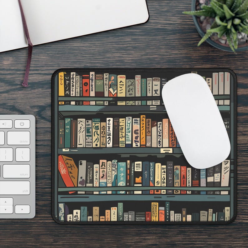 May include: A black mouse mat with a bookshelf design featuring rows of colourful books with Japanese text. The books are arranged on shelves with a dark wood finish. A white computer mouse is sitting on the mouse mat.