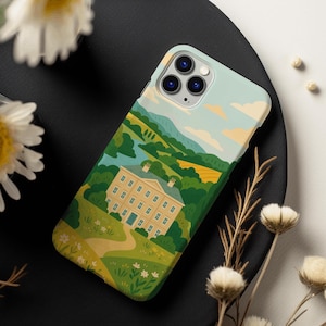May include: A phone case with a landscape design. It depicts a large yellow house with a blue door, set amidst green trees, fields, and a winding path. The sky is light blue with yellow clouds.
