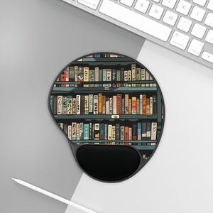 Peut inclure: Un tapis de souris ergonomique noir avec repose-poignet. Le tapis présente une illustration colorée d'une bibliothèque remplie de livres. Les livres ont du texte japonais sur leurs dos. Le tapis est sur une surface blanche près d'un clavier.