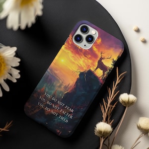 May include: A phone case with a sunset landscape and a deer standing on a cliff. The text "You do not fear, You do not falter, You do not yield" is printed on the case.