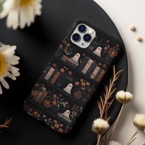Pode incluir: Uma capa de telefone preta com um design assustador que apresenta uma estante cheia de livros, flores e fantasmas. A capa tem um fundo escuro com detalhes laranja e branco.
