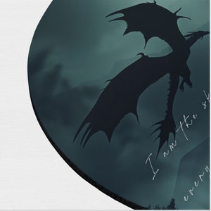 May include: A dark blue and black round coaster with a silhouette of a dragon flying in front of a cloudy sky. The text "I am the sky" is written in white on the coaster.