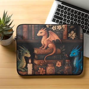 May include: A black laptop sleeve with a fantasy illustration of three dragons, books, and flowers. An orange dragon sits on books, with a green and blue dragon on either side. A candle and jars are also visible.