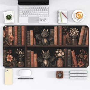 May include: A black desk mat featuring a bookshelf design with books, flowers, and two cartoon dragons. The mat has a white keyboard, mouse, and phone on the white surface. A cup of coffee and a notepad are also visible.