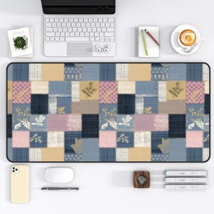 May include: A rectangular desk mat with a patchwork design in shades of blue, beige, and pink. The mat features a variety of floral and geometric patterns within the patchwork squares. The mat is surrounded by a black border and is placed on a white desk.