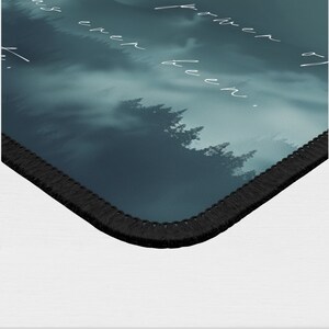 May include: A dark blue and green mousepad with a misty forest scene and the text "farther than ever been." The mousepad has a black stitched edge.
