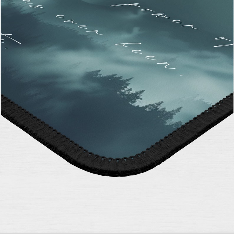 May include: A dark blue and green mousepad with a misty forest scene and the text "farther than ever been." The mousepad has a black stitched edge.