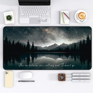 May include: A rectangular desk mat featuring a scenic landscape with a lake, mountains, and a starry night sky. The mat has the text "The mountains are calling and I must go." The mat is surrounded by a laptop, phone, and other office supplies.
