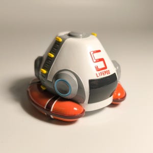 K&ouml;nnte beinhalten: Ein wei&szlig;-graues Modell einer Life Pod mit der Zahl 5 und dem Wort LIFE POD in Rot. Die Kapsel hat orangefarbene und braune Akzente und steht auf einer wei&szlig;en Oberfl&auml;che.