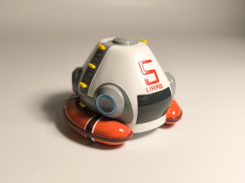 K&ouml;nnte beinhalten: Ein wei&szlig;-graues Modell einer Life Pod mit der Zahl 5 und dem Wort LIFE POD in Rot. Die Kapsel hat orangefarbene und braune Akzente und steht auf einer wei&szlig;en Oberfl&auml;che.