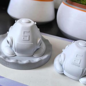 K&ouml;nnte beinhalten: Zwei hellgraue, futuristisch aussehende Objekte, m&ouml;glicherweise Masken oder Helme, werden gezeigt. Eines ruht auf einer grauen, kreisf&ouml;rmigen Basis, w&auml;hrend das andere direkt auf einer Oberfl&auml;che liegt. Jedes hat die Zahl "5" und andere Markierungen.