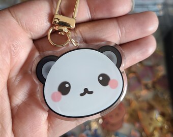 Könnte beinhalten: Ein klarer Acryl-Panda-Schlüsselanhänger mit einem goldfarbenen Verschluss. Der Panda hat ein weißes Gesicht, schwarze Ohren und rosa Wangen. Der Schlüsselanhänger wird in einer Hand gehalten.