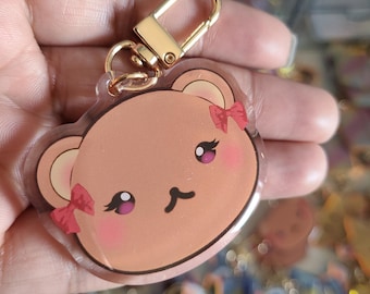Könnte beinhalten: Ein klarer Acryl-Schlüsselanhänger mit einem Cartoon-Bärengesicht. Der Bär hat rosa Wangen, kleine rote Schleifen und goldfarbene Hardware. Das Gesicht des Bären hat dunkle Augen und eine kleine schwarze Nase. Der Schlüsselanhänger wird in einer Hand gehalten.