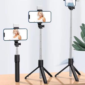May include: Three black and silver selfie sticks with phones attached, each displaying a woman. The selfie sticks have adjustable heights and tripod bases. Each phone has a white light attached.