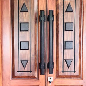 Puede incluir: Un par de puertas de madera con acabado marrón rojizo. Cada puerta presenta un diseño decorativo con formas geométricas negras, incluyendo triángulos y cuadrados, y manijas verticales negras. Una llave está en la cerradura.
