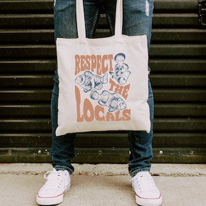 May include: A white canvas tote bag with the text "Respect the Locals" in orange and blue lettering. The bag features a graphic of three fish with blue and white details.