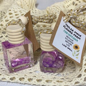 May include: Two car air fresheners with purple liquid and wooden tops. Each has a tag that reads "Taylor Made Flower Farm Car Diffuser" and "Love Spell". The bottles are on a cream-colored, open-weave surface.