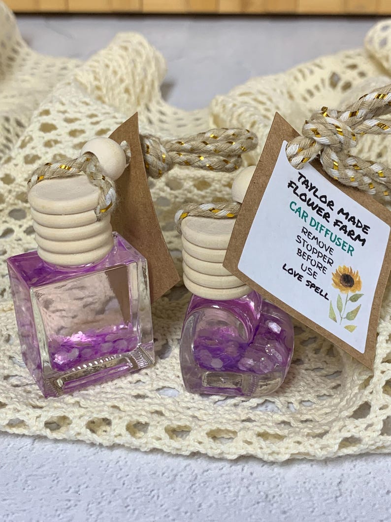 May include: Two car air fresheners with purple liquid and wooden tops. Each has a tag that reads "Taylor Made Flower Farm Car Diffuser" and "Love Spell". The bottles are on a cream-colored, open-weave surface.