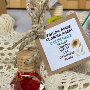 May include: A car air freshener with a red, diamond-shaped glass bottle and a wooden cap. The bottle is suspended by a beige and gold rope. A brown tag reads "Taylor Made Flower Farm Car Diffuser" and "Cashmere Plum".