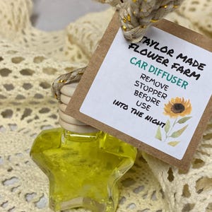 May include: A star-shaped car air freshener filled with yellow liquid, hanging from a tag with the text "Taylor Made Flower Farm Car Diffuser." The tag also features a sunflower illustration. The freshener is attached to a beige rope with gold accents.
