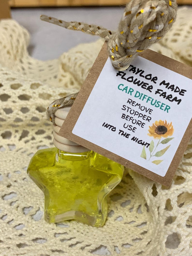 May include: A star-shaped car air freshener filled with yellow liquid, hanging from a tag with the text "Taylor Made Flower Farm Car Diffuser." The tag also features a sunflower illustration. The freshener is attached to a beige rope with gold accents.