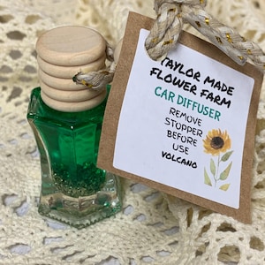 May include: A car air freshener with a green liquid inside a clear, geometric-shaped glass bottle. The bottle has a wooden stopper and is attached to a tag with the text "Taylor Made Flower Farm Car Diffuser". A sunflower illustration is also on the tag.