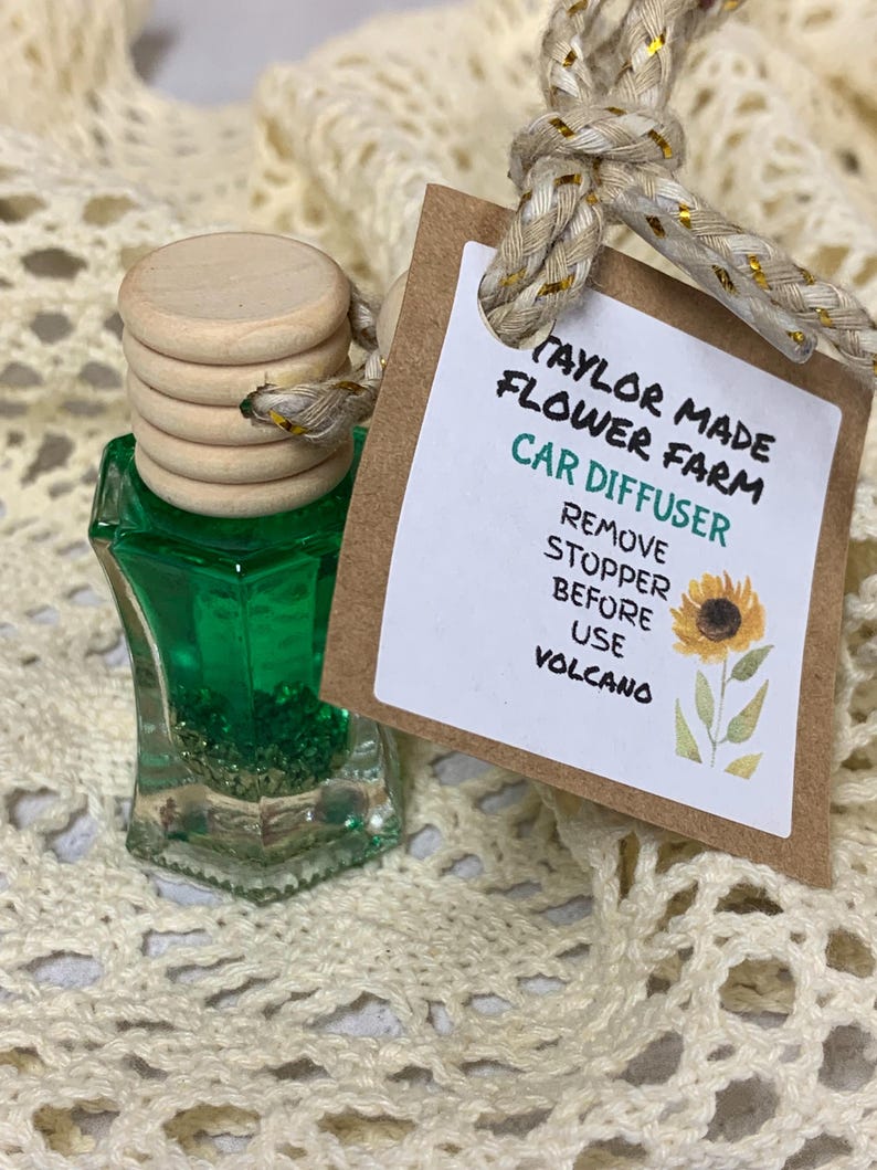 May include: A car air freshener with a green liquid inside a clear, geometric-shaped glass bottle. The bottle has a wooden stopper and is attached to a tag with the text "Taylor Made Flower Farm Car Diffuser". A sunflower illustration is also on the tag.