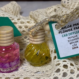 May include: Two car air fresheners with wooden tops and colorful liquid. One is pink and purple, the other is yellow. A tag reads "Taylor Made Flower Farm Car Diffuser" with instructions and a sunflower illustration.