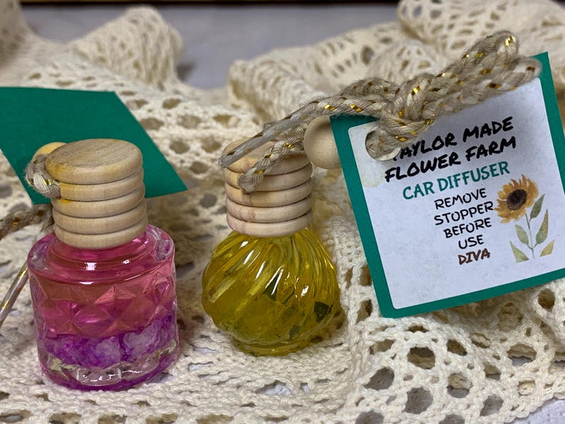 May include: Two car air fresheners with wooden tops and colorful liquid. One is pink and purple, the other is yellow. A tag reads "Taylor Made Flower Farm Car Diffuser" with instructions and a sunflower illustration.