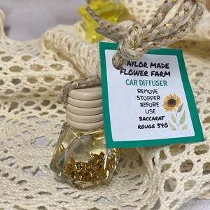 May include: A clear, geometric-shaped car air freshener with a wooden cap and a decorative rope. The label reads "Taylor Made Flower Farm Car Diffuser" and includes instructions. The bottle contains gold-colored beads and is displayed on a cream-colored, open-weave fabric.