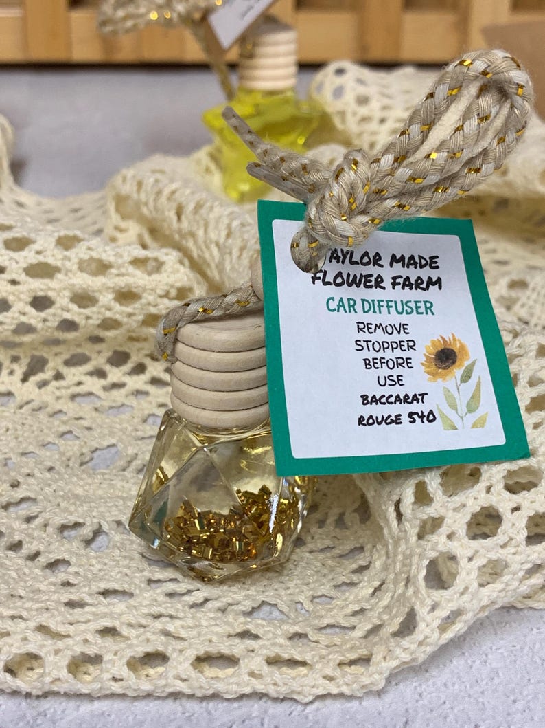 May include: A clear, geometric-shaped car air freshener with a wooden cap and a decorative rope. The label reads "Taylor Made Flower Farm Car Diffuser" and includes instructions. The bottle contains gold-colored beads and is displayed on a cream-colored, open-weave fabric.
