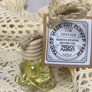 May include: A clear glass car air freshener bottle with a wooden top and a decorative cord. The bottle contains a yellow liquid and is labeled "Made Cut Flower Car Diffuser French Market". The cord is beige with gold accents.