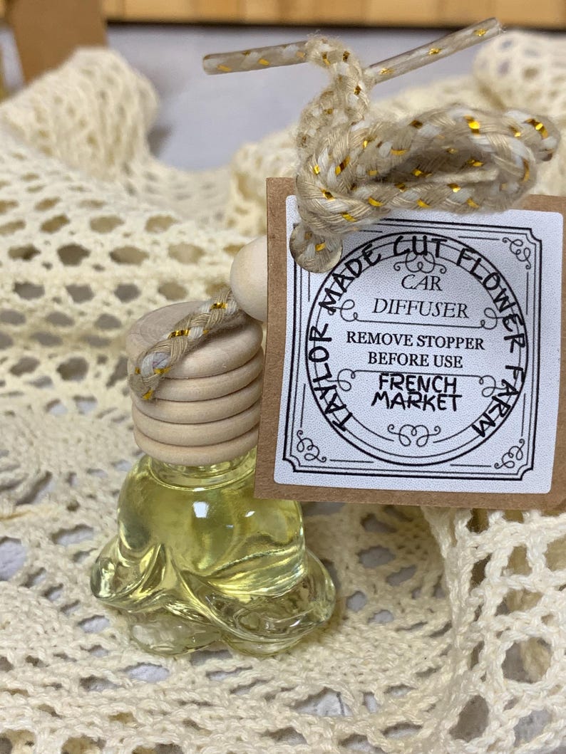 May include: A clear glass car air freshener bottle with a wooden top and a decorative cord. The bottle contains a yellow liquid and is labeled "Made Cut Flower Car Diffuser French Market". The cord is beige with gold accents.