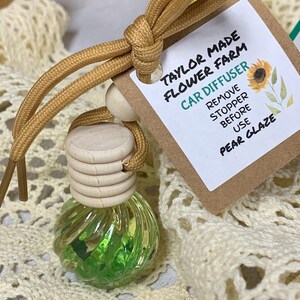 May include: A car air freshener with a pear glaze scent. The glass bottle is filled with green liquid and decorative elements. A tag reads "Taylor Made Flower Farm Car Diffuser" with instructions. The freshener is attached to a tan cord.