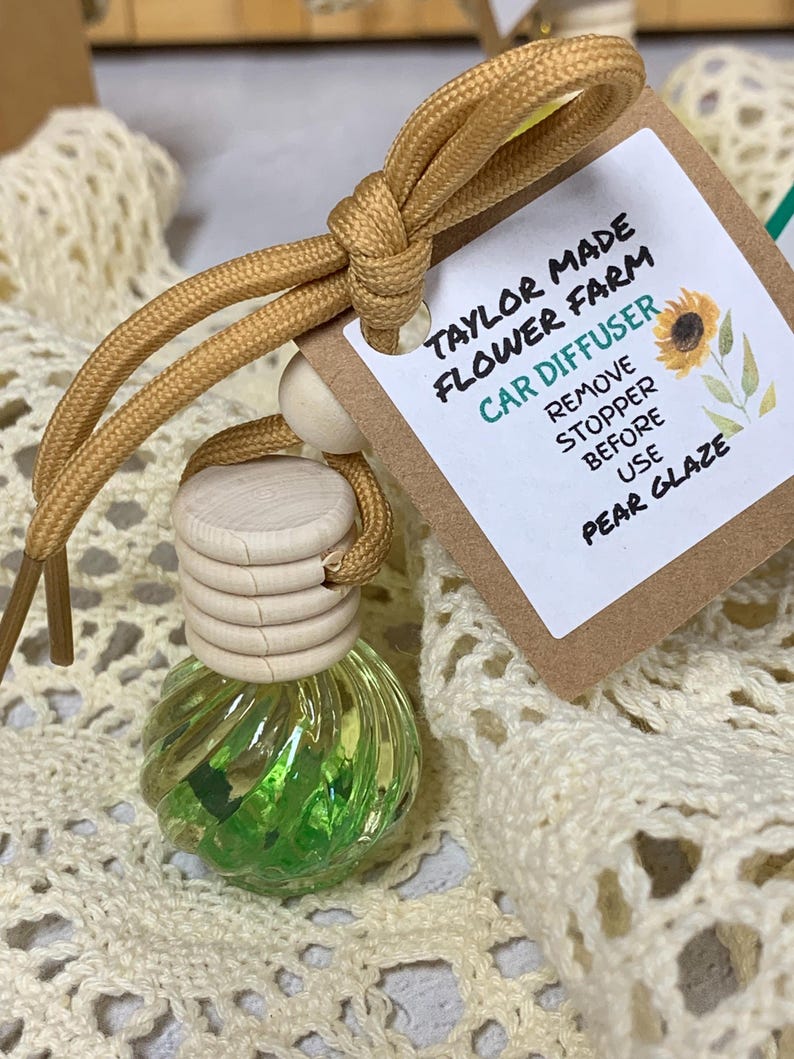 May include: A car air freshener with a pear glaze scent. The glass bottle is filled with green liquid and decorative elements. A tag reads "Taylor Made Flower Farm Car Diffuser" with instructions. The freshener is attached to a tan cord.