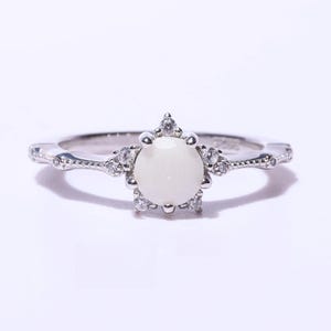 May include: A silver ring with a large, round, off-white stone at the center, surrounded by smaller clear stones. The band has a delicate, ornate design with additional small stones.