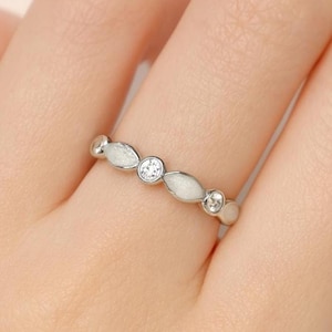 May include: A silver ring with a repeating pattern of white, oval-shaped accents and clear, round gemstones. The ring is worn on a finger, showcasing its delicate design and sparkling details. The ring's design is elegant and suitable for everyday wear.