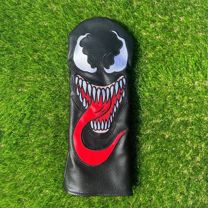 K&ouml;nnte beinhalten: Schwarzer Golfschl&auml;gerkopfbezug mit einem roten und wei&szlig;en Venom-Charakterdesign. Der Charakter hat einen gro&szlig;en Mund mit scharfen Z&auml;hnen und eine lange rote Zunge.