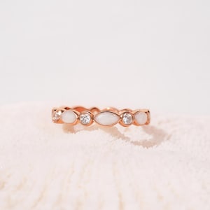 May include: Rose gold ring featuring alternating white oval and round clear stones. The ring has a delicate design, with the stones set closely together. The ring is displayed on a white, textured surface.