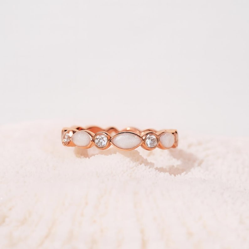 May include: Rose gold ring featuring alternating white oval and round clear stones. The ring has a delicate design, with the stones set closely together. The ring is displayed on a white, textured surface.