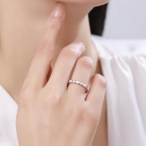May include: A silver ring with a delicate design featuring alternating oval and round white gemstones. The ring is worn on a finger, showcasing its elegant and minimalist style. The ring is a piece of jewelry.