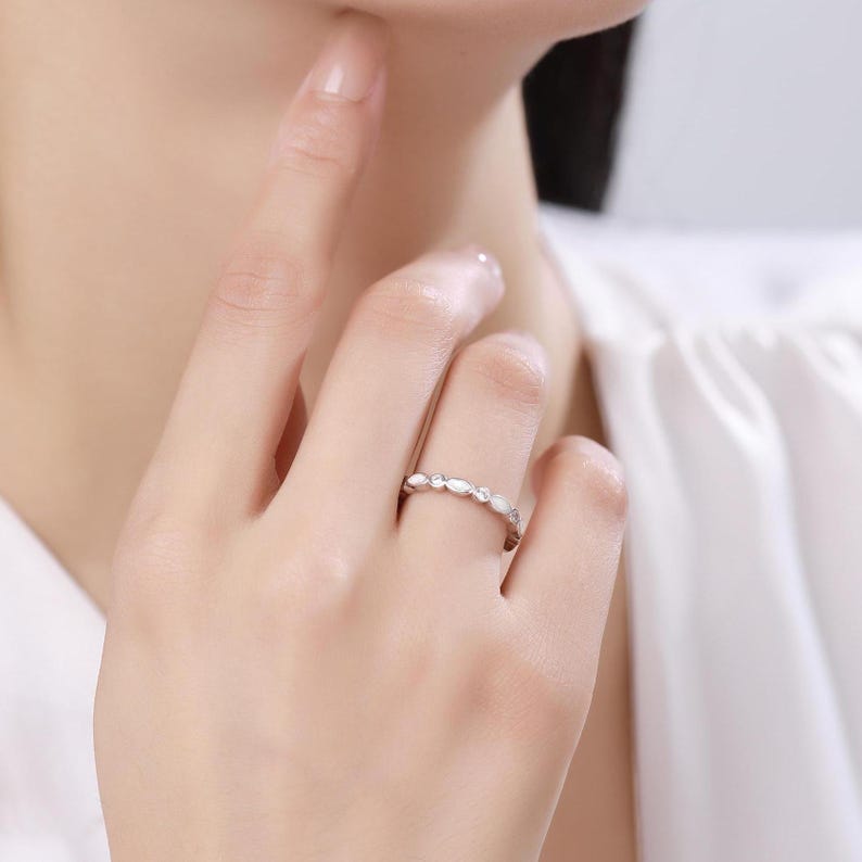 May include: A silver ring with a delicate design featuring alternating oval and round white gemstones. The ring is worn on a finger, showcasing its elegant and minimalist style. The ring is a piece of jewelry.