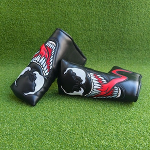 Könnte beinhalten: Zwei schwarze Golfschlägerhauben mit gestickten Designs. Eine Haube zeigt eine rote Zunge und weiße Zähne, während die andere ein ähnliches Design hat. Die Hauben liegen auf einer grünen Oberfläche.