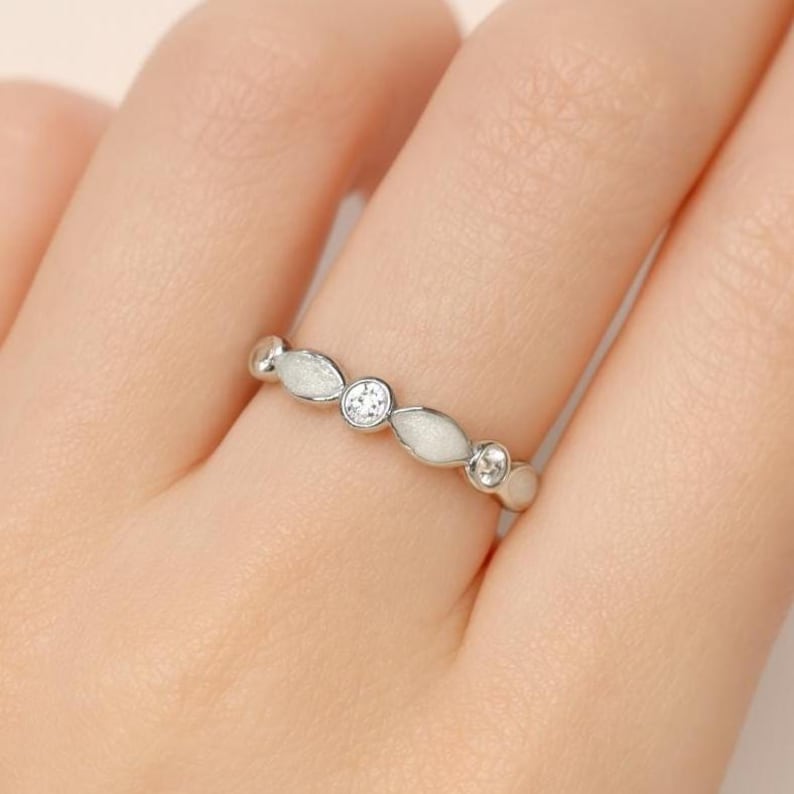 May include: A silver ring with a repeating pattern of white, oval-shaped accents and clear, round gemstones. The ring is worn on a finger, showcasing its delicate design and sparkling details. The ring's design is elegant and suitable for everyday wear.