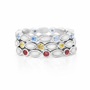 May include: Three silver rings stacked together, each featuring a repeating pattern of white enamel and colorful gemstones. The top ring has blue gems, the middle has yellow, and the bottom has red.