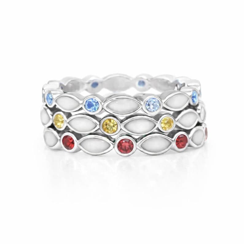 May include: Three silver rings stacked together, each featuring a repeating pattern of white enamel and colorful gemstones. The top ring has blue gems, the middle has yellow, and the bottom has red.